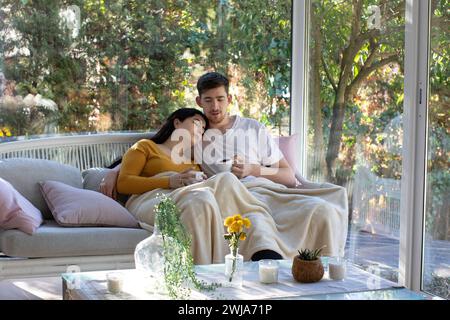 Young man and woman with cups of coffee sitting on sofa covering legs with blanket while spending time together on weekend in modern living room Stock Photo