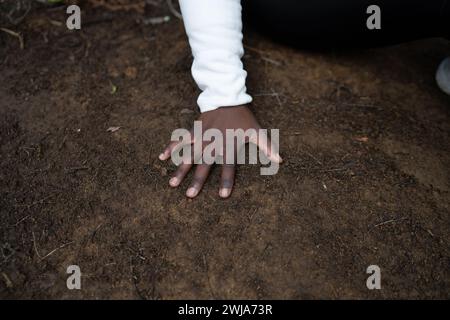 From above of anonymous ethnic female in full sleeve jacket while in dark area placing open palm hand on wet soil ground in light Stock Photo