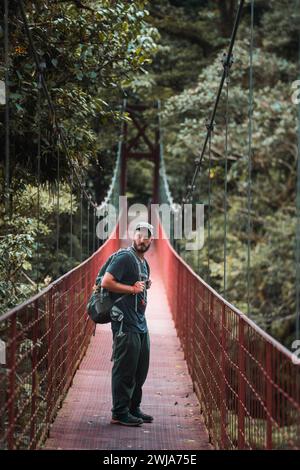 A male hiker with a backpack stands midway on a red suspension bridge surrounded by dense vegetation, looking pensively at the camera. Stock Photo