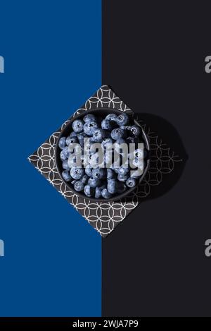 Top view of bowl of juicy blueberries on a geometric patterned napkin, set against a contrasting blue and black diagonal background Stock Photo