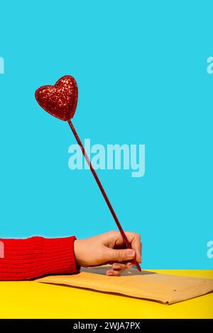 Unrecognizable female in full sleeve sweater and with manicured fingers writing on envelope cover on yellow surface with glittering heart shaped mock Stock Photo