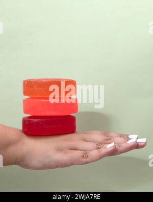 A hand presented against a neutral background balances three colorful bars of natural homemade soap, showcasing shades of orange and red. Stock Photo