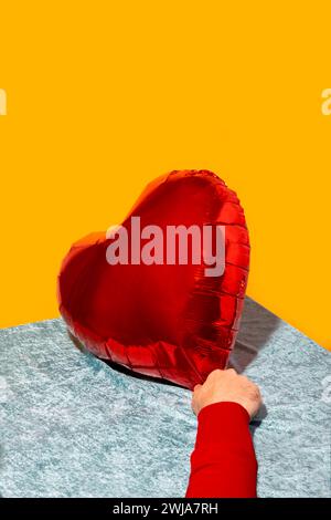 From above of unrecognizable person hand in full sleeve sweater placing big bright red heart shaped balloon on floral fabric surface against yellow ba Stock Photo