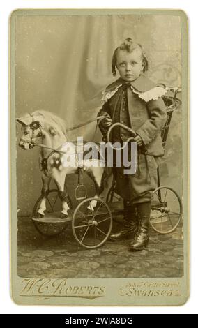 Original, charming, Victorian CDV  Carte de Visite ( visiting card or CDV) of cute young Welsh Victorian boy, Victorian child, with hair in ringlets, wearing an elaborate Little Lord Fauntleroy outfit, with a lace collar, knickerbockers probably the studio's as is outsize!, standing next to a beaurtiful toy horse on wheels, a studio prop, posing for a  portrait at the photographic studio of W C Roberts of 16 17 Castle St. Swansea, U.K. circa late 1880's - 1890's. Stock Photo