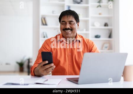 Positive middle aged hindu businessman using smartphone and laptop Stock Photo