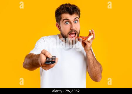 guy points remote controller savoring pizza slice on yellow backdrop Stock Photo