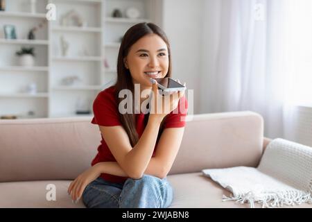 Portrait Of Smiling Young Asian Woman Recording Voice Message On Smartphone Stock Photo