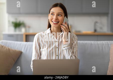 Happy young woman talking on phone and using laptop at home Stock Photo
