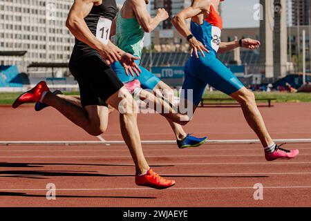 three male athletes sprinting on red track, muscles taut, competing fiercely. Brightly colored shoes emphasize motion Stock Photo