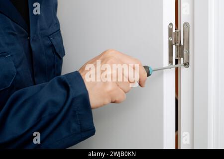 a caucasian man, in blue overalls, uses a screwdriver to screw or unscrew the hinge of a white indoors door Stock Photo