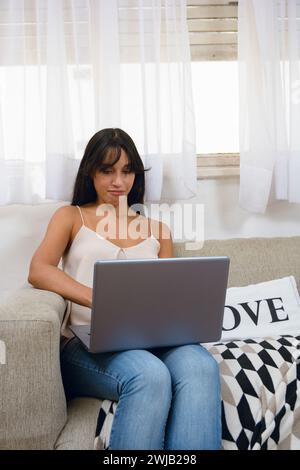 vertical image, young Hispanic Latina entrepreneur woman at home happy sitting on couch working with her laptop, business concept, copy space. Stock Photo