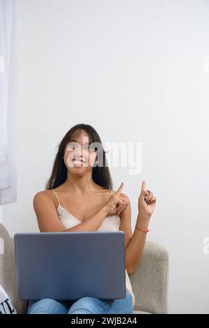 young enterprising brunette latina woman over white wall at home with her laptop pointing up and looking at camera smiling with laptop on her legs and Stock Photo