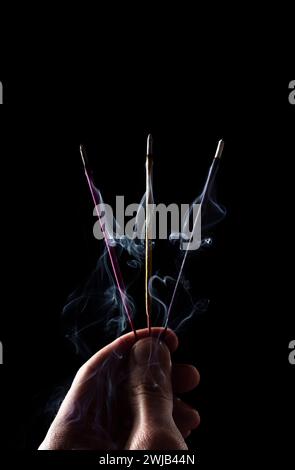 Three multi-colored incense sticks in a man's hand. Sticks are smoking on a black background. Stock Photo