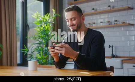 Man playing mobile game on the smartphone while sitting in kitchen at home. Excited man engrossed in interesting game, doing winner gesture Stock Photo