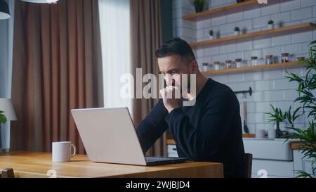 Frustrated man receives bad news sees shocking information on laptop screen while sitting in kitchen at home reacts with emotions. Upset man feeling s Stock Photo