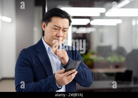 Man in business suit inside office with phone in hands, mature businessman received online notification message with bad news, boss uses app on smartphone, upset received rejection error. Stock Photo