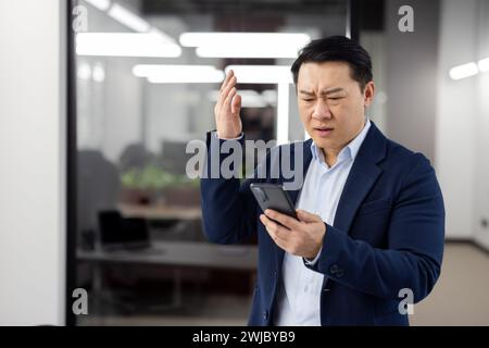 Man in business suit inside office with phone in hands, mature businessman received online notification message with bad news, boss uses app on smartphone, upset received rejection error. Stock Photo