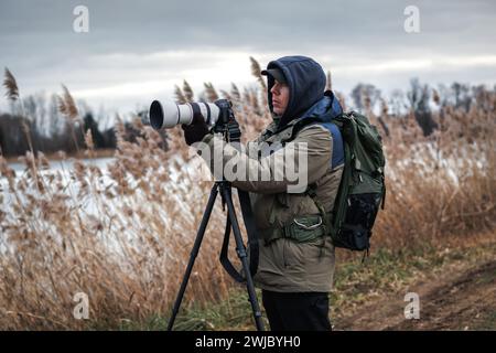 Photographer is setting up camera on tripod outdoors. Man photographing landscape or wildlife at lake in winter Stock Photo