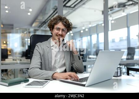 Smiling young businessman with curly hair focused on work at his laptop in a well-lit modern corporate office setting. Stock Photo