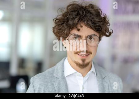Portrait of a confident young businessman with curly hair, wearing glasses, and smiling in a bright office environment. Stock Photo