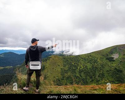 A hiker standing on a hilltop points his hand towards the fog-covered mountains Stock Photo