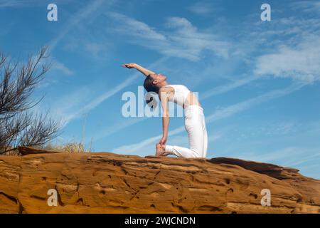 fit woman doing a camel yoga pose on red rocks, blue sky background Stock Photo