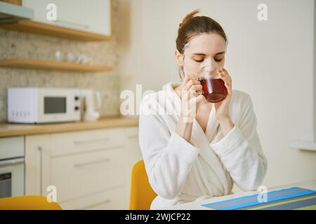 A woman with a cosmetic anti-aging mask on her face sits in the kitchen and drinks tea Stock Photo