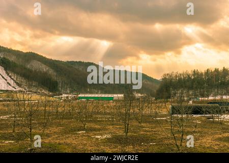 Sun rays break through dark clouds sending beans of light to wintery rural landscape below Stock Photo
