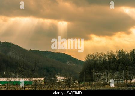 Sun rays break through dark clouds sending beans of light to wintery rural landscape below Stock Photo