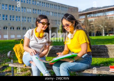 Multi-ethnic students with headphones looking at homework sitting on the bench of a campus Stock Photo