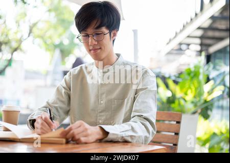 A happy Asian man is focusing on writing in his notebook, jotting down ideas, or keeping a diary while sitting at an outdoor table in a cafe in the ci Stock Photo