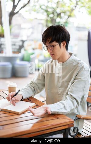 A happy Asian man is focusing on writing in his notebook, jotting down ideas, or keeping a diary while sitting at an outdoor table in a cafe in the ci Stock Photo