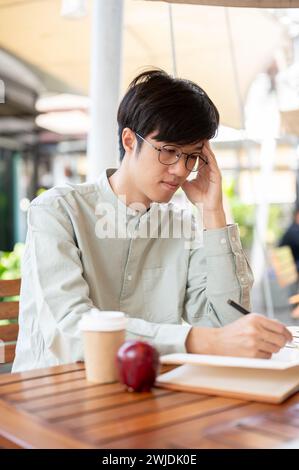 A focused and serious young Asian man is contemplating and jotting down his ideas in a book while sitting at an outdoor table in a cafe. people and li Stock Photo