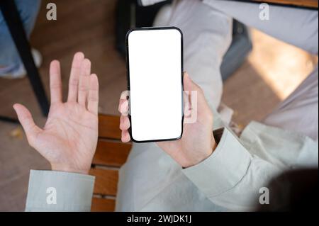 A top view image of an unhappy, dissatisfied man showing his palm while using his smartphone at a table indoors. A white-screen smartphone mockup. Stock Photo