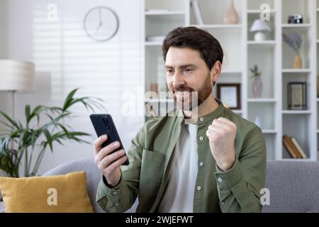Bearded mature man showing joy while using his smartphone on a couch. Success and happiness at home depicted in modern living room. Stock Photo