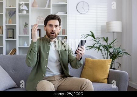 Adult man expressing shock and confusion while looking at his smartphone, seated comfortably on a living room sofa. Stock Photo