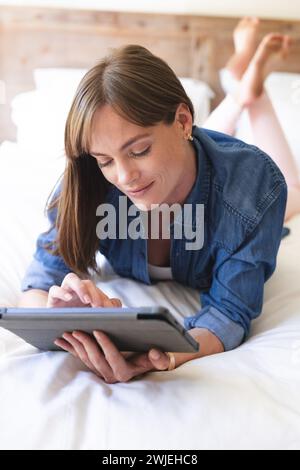 A young Caucasian woman is focused on her tablet while lying on a bed Stock Photo
