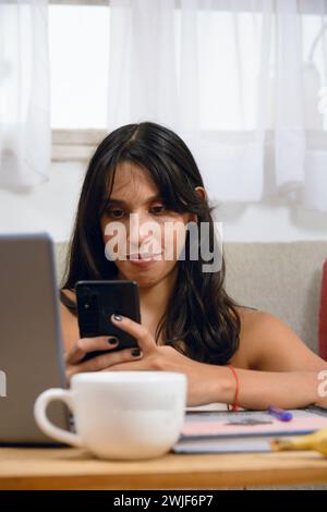 vertical portrait of young brunette latin woman student at home attentive watching her phone sitting on floor with her laptop on table, copy space. Stock Photo