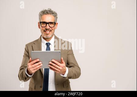 A cheerful senior businessman confidently stands with a tablet in hands, indicating the importance of digital devices in the business landscape and executive accessibility. Stock Photo