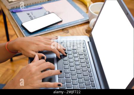 Close-up of female brunette hands of unrecognizable person with black painted nails, typing on laptop keyboard on table at home next to phone and note Stock Photo
