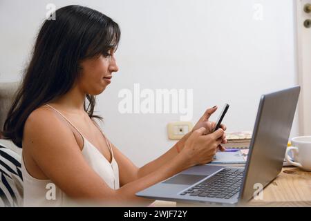 side view of young brunette latina woman at home sitting on floor, working with laptop and checking cell phone checking message from her clients. copy Stock Photo