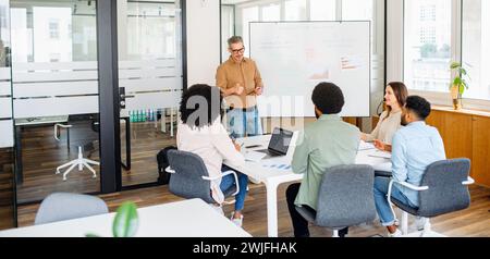 A charismatic mature male presenter leads a dynamic brainstorming session in a contemporary office setting, diverse team listens intently. A vibrant exchange of ideas and a culture of collaboration Stock Photo