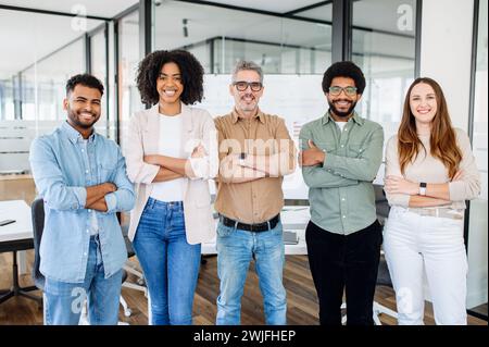A dynamic team of five, radiating positivity and diversity, stand confidently in co-working space, the seasoned team leader at center, exemplifying unity and successful collaboration in the workplace. Stock Photo