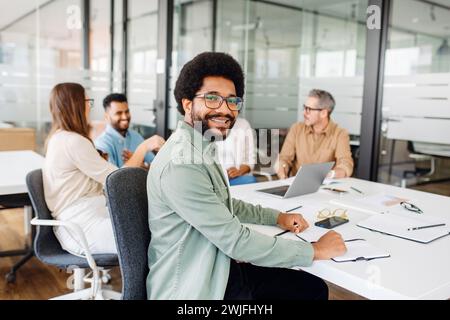 Brazilian man in a green shirt and glasses is seated at a modern office table and smiling at the camera, while colleagues converse in the background, depicting a casual yet focused working atmosphere Stock Photo