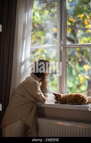 Melancholic pet owner looking at window in depression while staying at home in sunny summer day Stock Photo