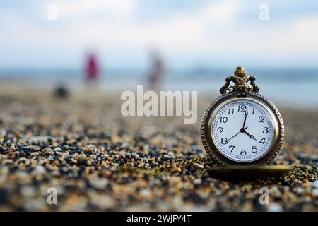Gold pocket watch lies on a sandy beach with a person walking on the beach in the background. With copy space for text Stock Photo