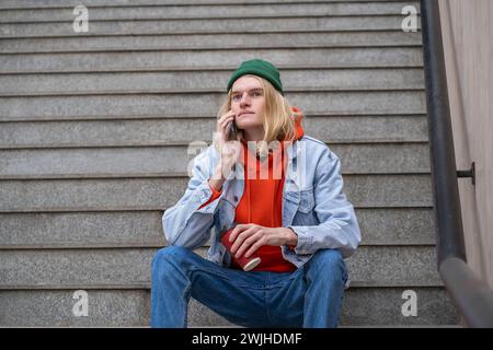 Hipster blond guy with coffee cup in hand sits on steps talks on smartphone in city street.  Stock Photo