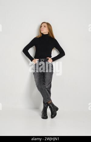 A trendy young woman poses confidently, wearing a fitted black top, grey jeans, and black boots against a white background Stock Photo