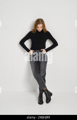 A young woman wearing a fitted black sweater, grey jeans, and black boots leaning against a white wall and looks down Stock Photo