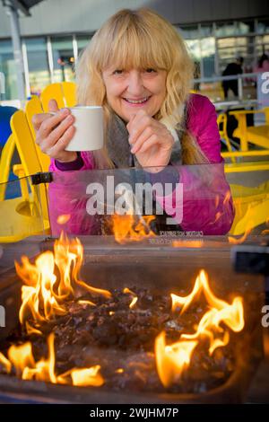 Woman enjoying mug of coffee outdoors by warm fire in winter Stock Photo